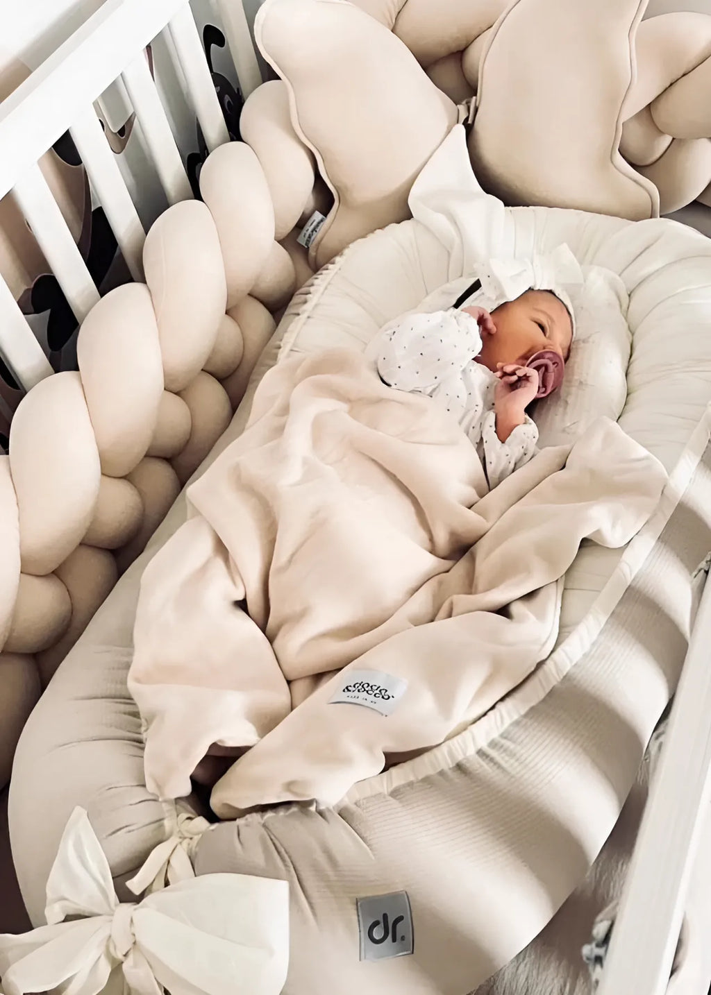newborn in dockatot surrounded by cream 4 strand braided crib bumper, white standard crib, safe for newborns