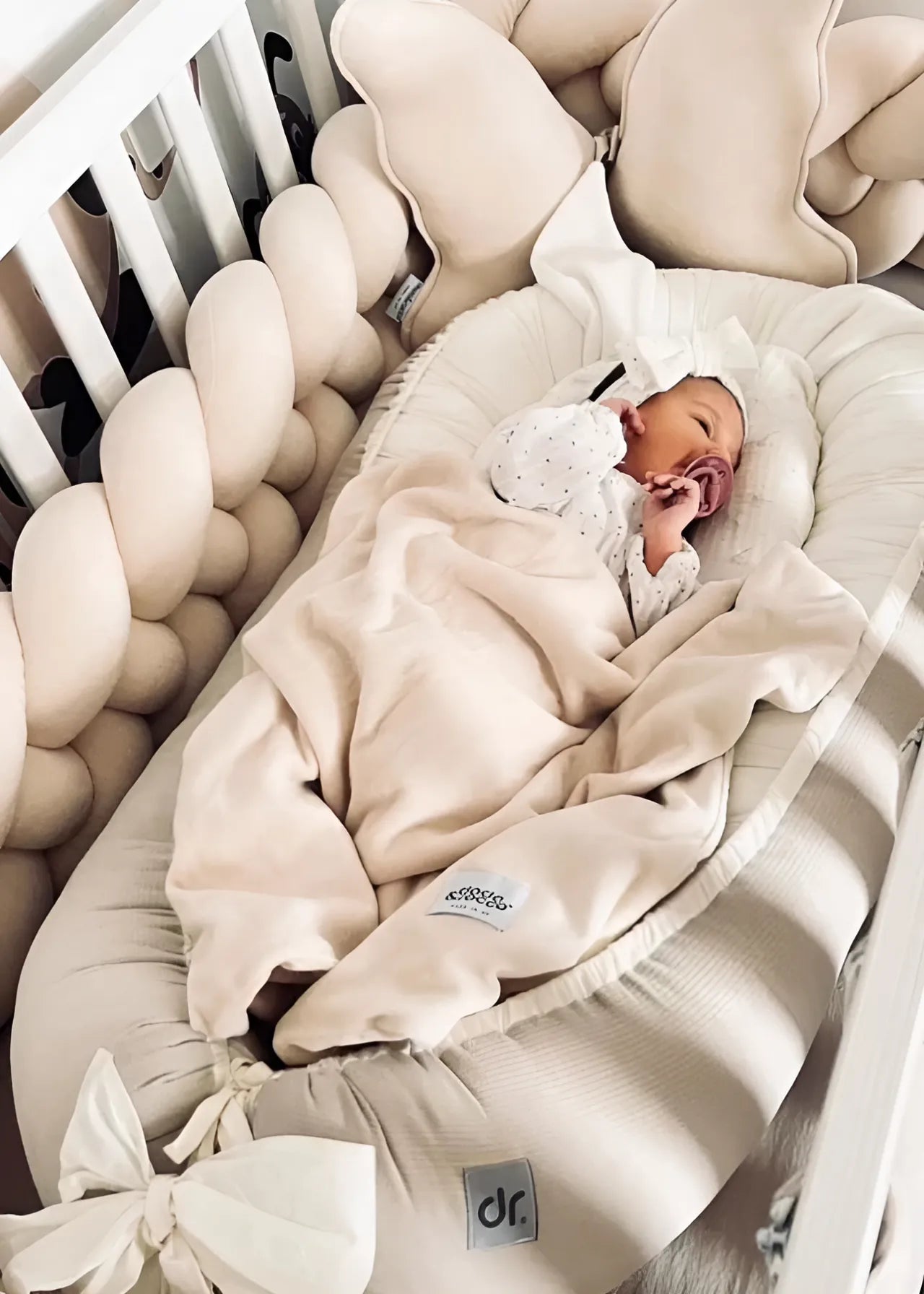 newborn in dockatot surrounded by cream 4 strand braided crib bumper, white standard crib, safe for newborns