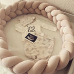 Cream 3-strand braided crib bumper arranged in circle with newborn clothes and ultrasound, pregnancy announcement